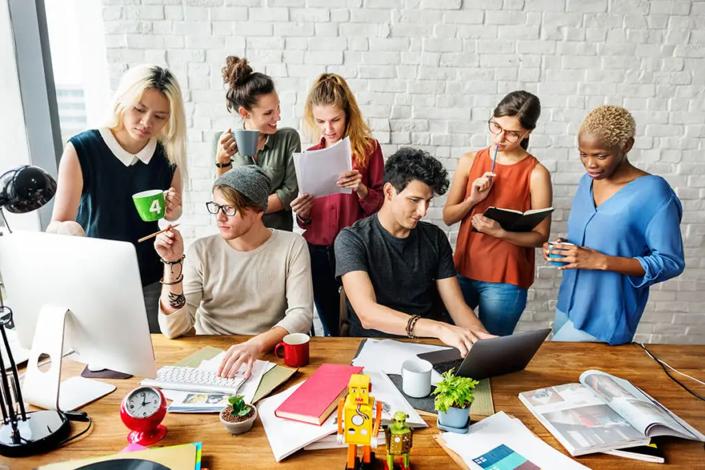 une équipe de collaborateurs assis derrière une table de bureau en bois, avec un mur en briques blanc en arrière plan, courtier assurance vie luxembourg